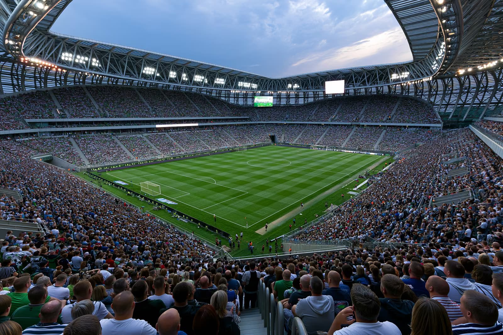 Një stadium i madh futbolli i mbushur me tifozë nën dritat e prozhektorëve në një mbrëmje vere.