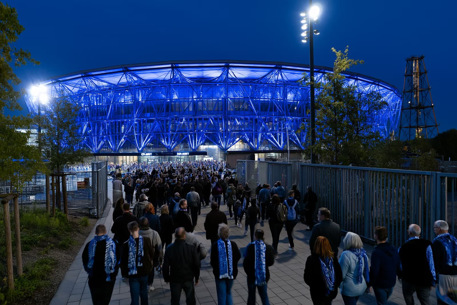 Një stadium futbolli në zonën e Ruhr-it shkëlqen me blu mbretërore natën, ndërsa tifozët me shalle vërshojnë drejt portave pas transferimit të Gosens Schalke.