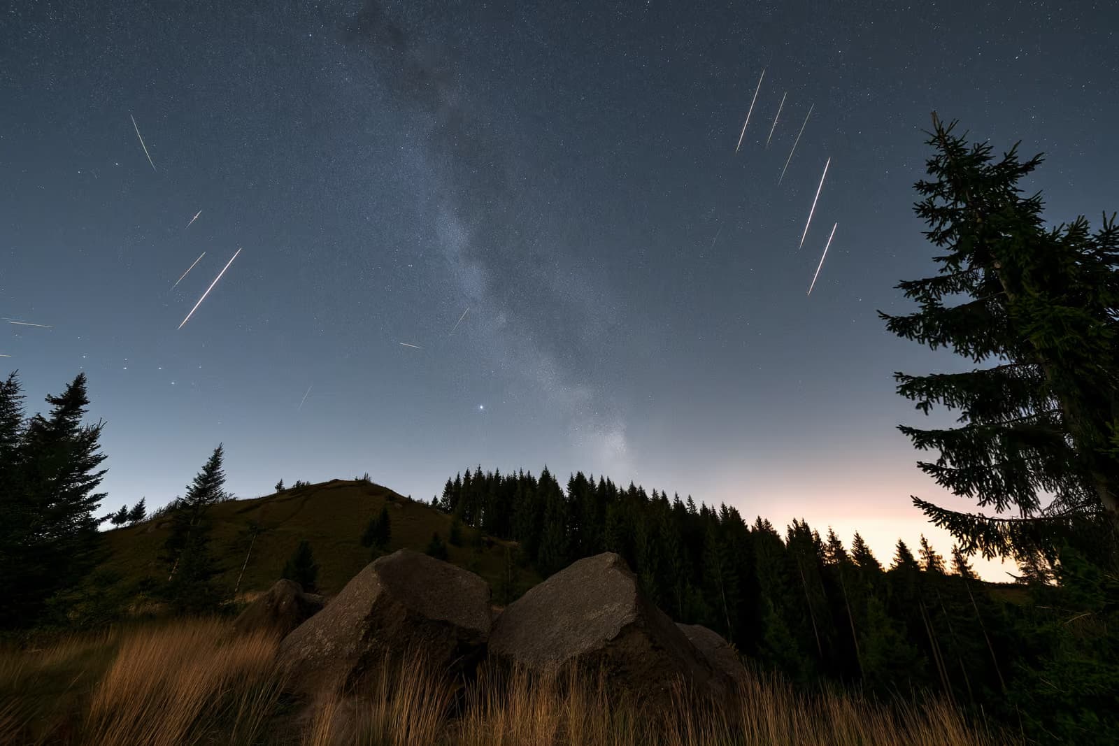 Një qiell nate mbi një fshat të errët gjerman me vija meteorësh dhe një brez të zbehtë yjesh sipër kokës.
